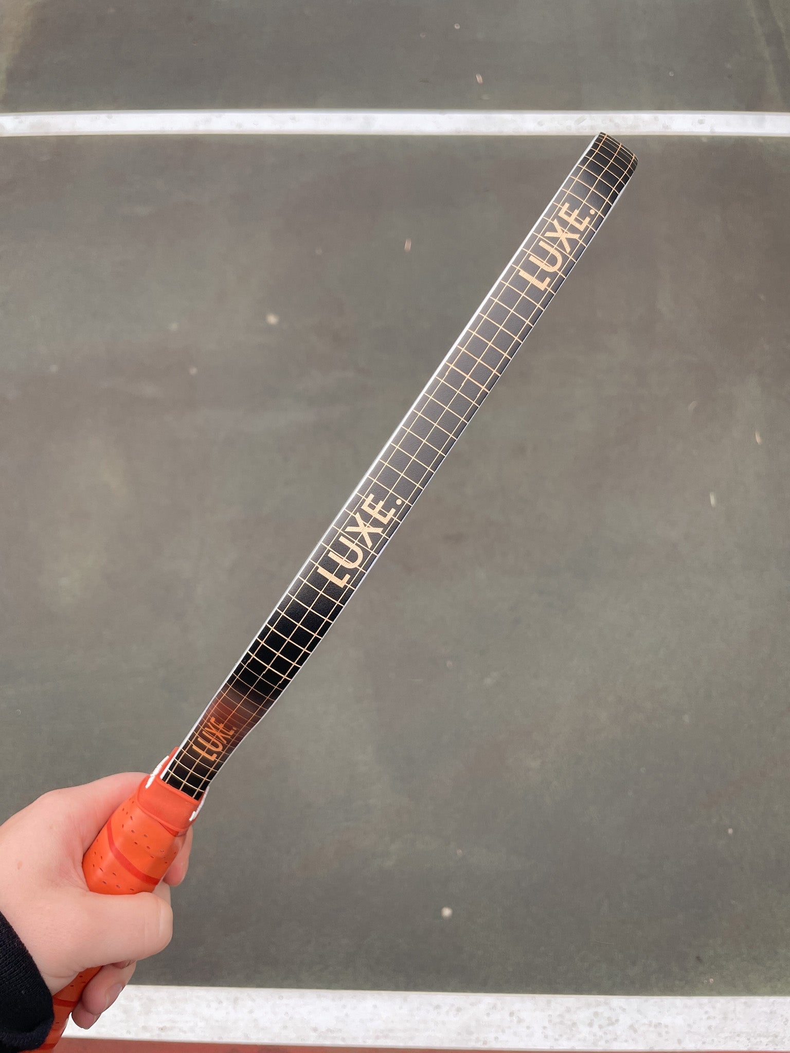 Hand holding a tennis racquet with a black and tan checkered pattern and the word "LUXE" printed on it.