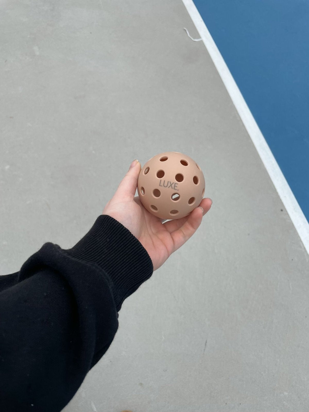 Person holding a perforated pink pickleball with the word "LUXE" on it in their hand while wearing a black sweatshirt. Background of gray concrete with a white line and blue painted area.