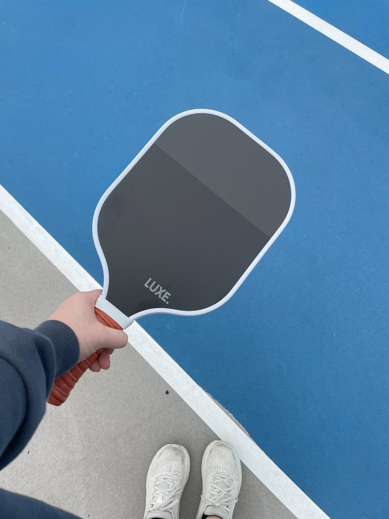 Person holding a gray and white pickleball paddle with an orange grip, wearing white sneakers on a blue and gray court.