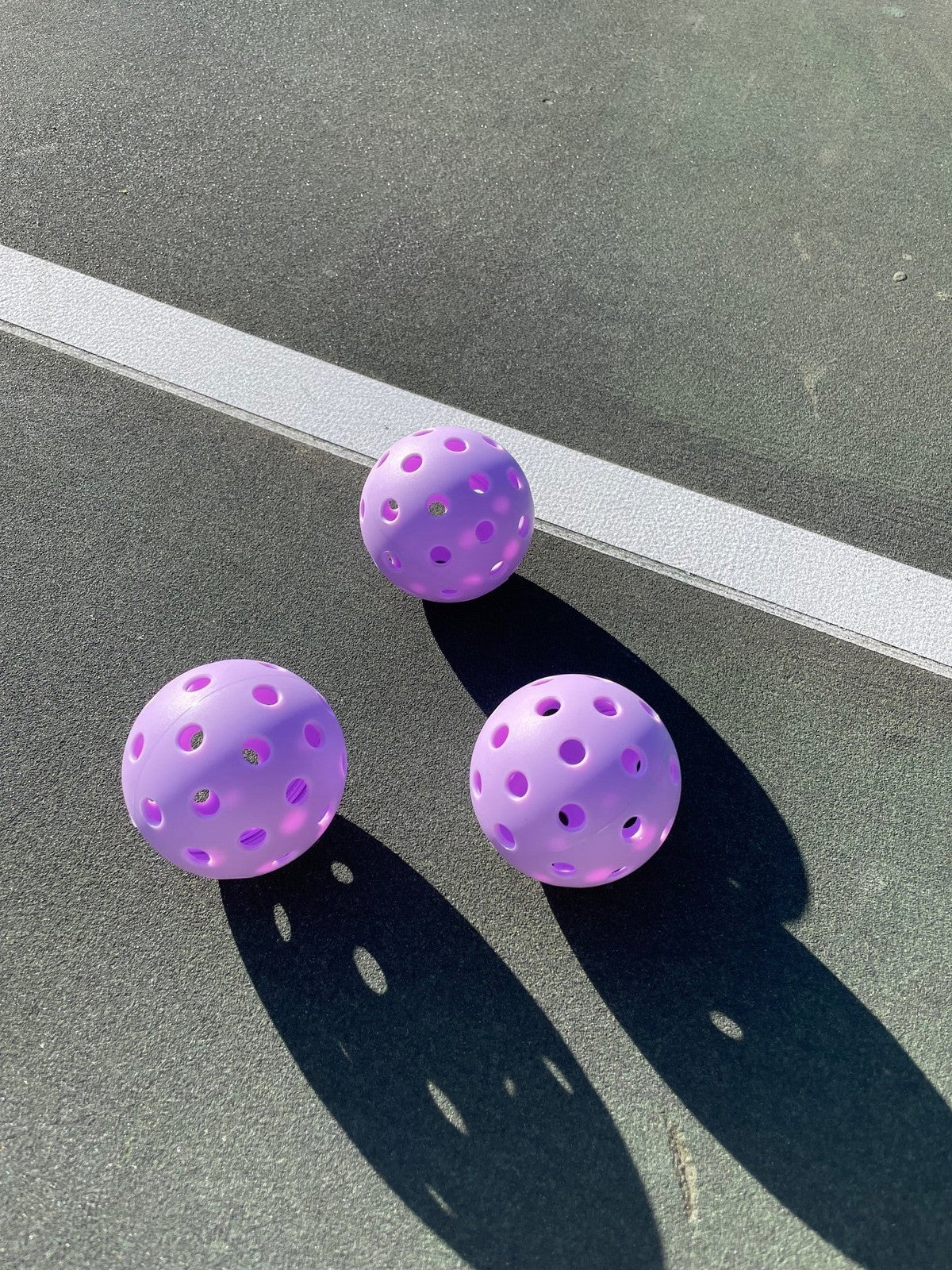 Three purple pickleballs cast shadows on a dark green court surface near a white line.