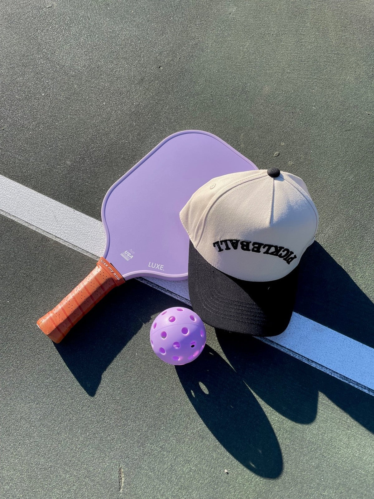 A pickleball paddle, ball, and baseball cap are arranged on a green court with a white line. The paddle is light purple with a brown grip, the ball is also light purple with holes, and the cap has a tan crown and a black brim with "PICKLEBALL" stitched on it in black.