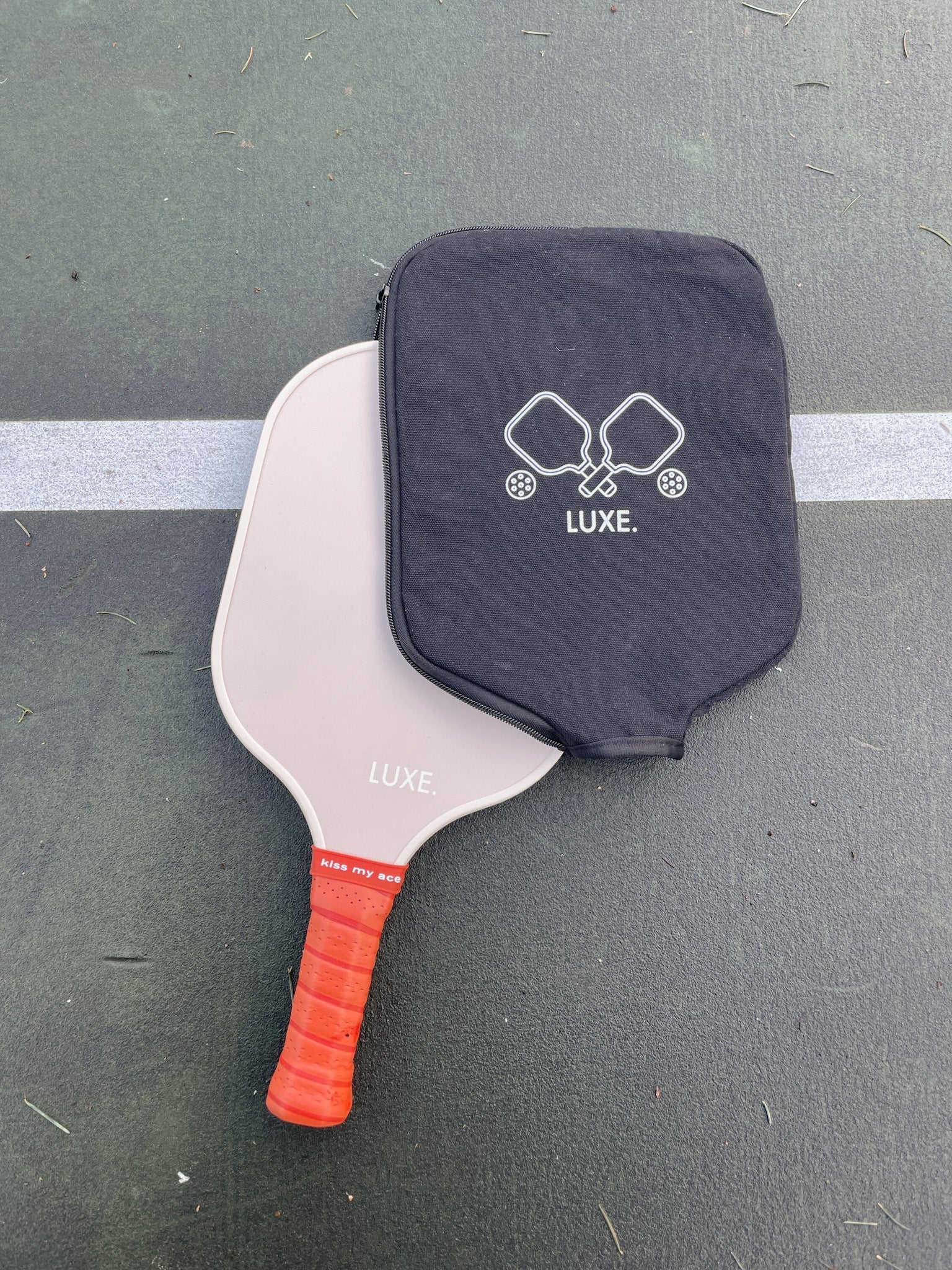 Overhead shot of a pickleball paddle with an orange grip and the words "kiss my ace," next to a black zippered case with a logo that reads "LUXE." The objects are laying on a green pickleball court with a white line.