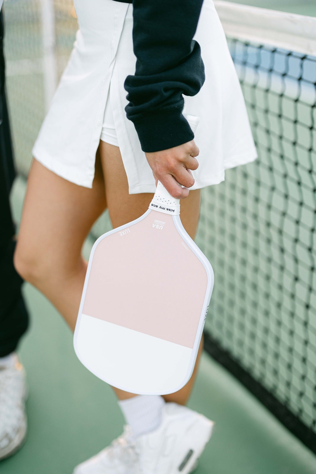A woman in a white tennis skirt and black sweatshirt holds a pink and white pickleball paddle.