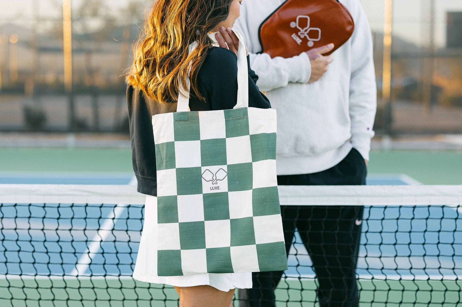 Two people at a pickleball court. One is holding a green and white checkered tote bag. The other is holding a pickleball paddle case.