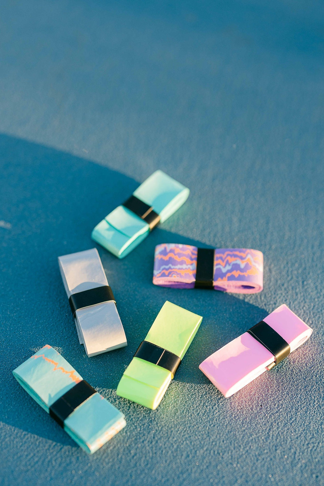 A high-angle shot shows five colorful rolls of athletic tape on a textured blue surface. Each roll is secured with a black band around the center. The colors of the tape include aqua, gray, purple with a wavy pattern, bright green, and pink.