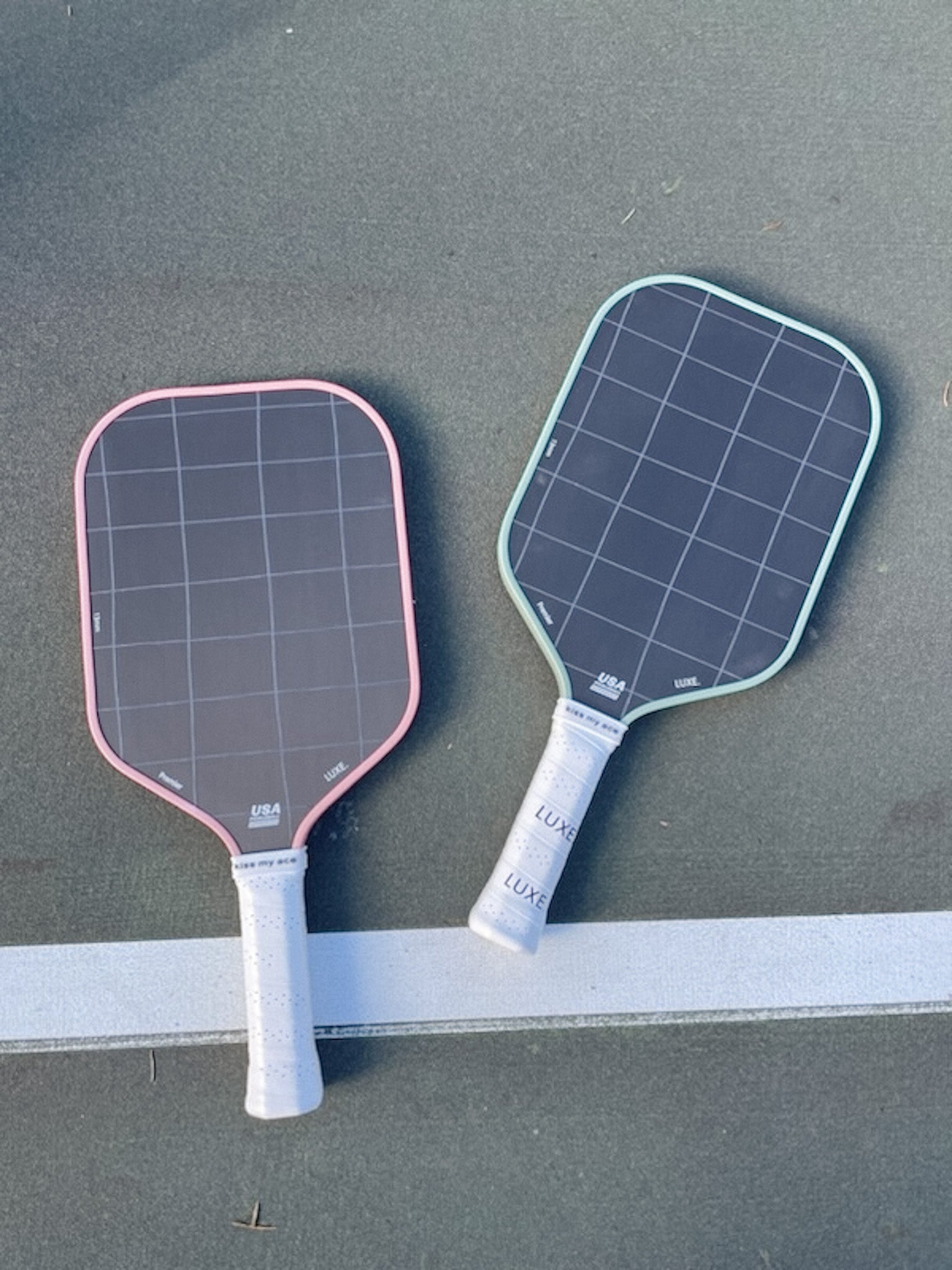 Two pickleball paddles, one with a pink edge and one with a light blue edge, lying on a grey surface with a white line.
