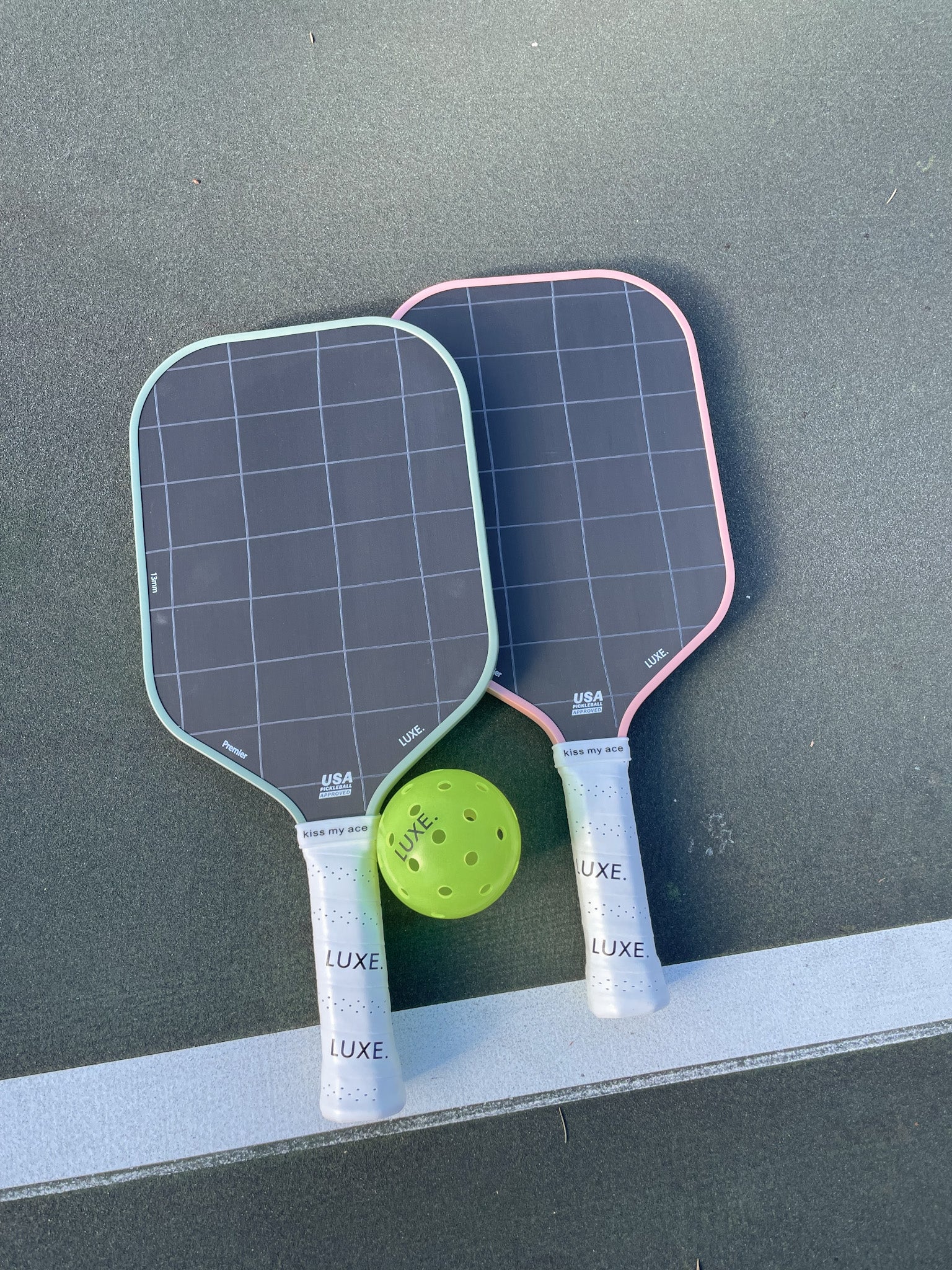 Two pickleball paddles with a green pickleball ball on a green surface with a white line.