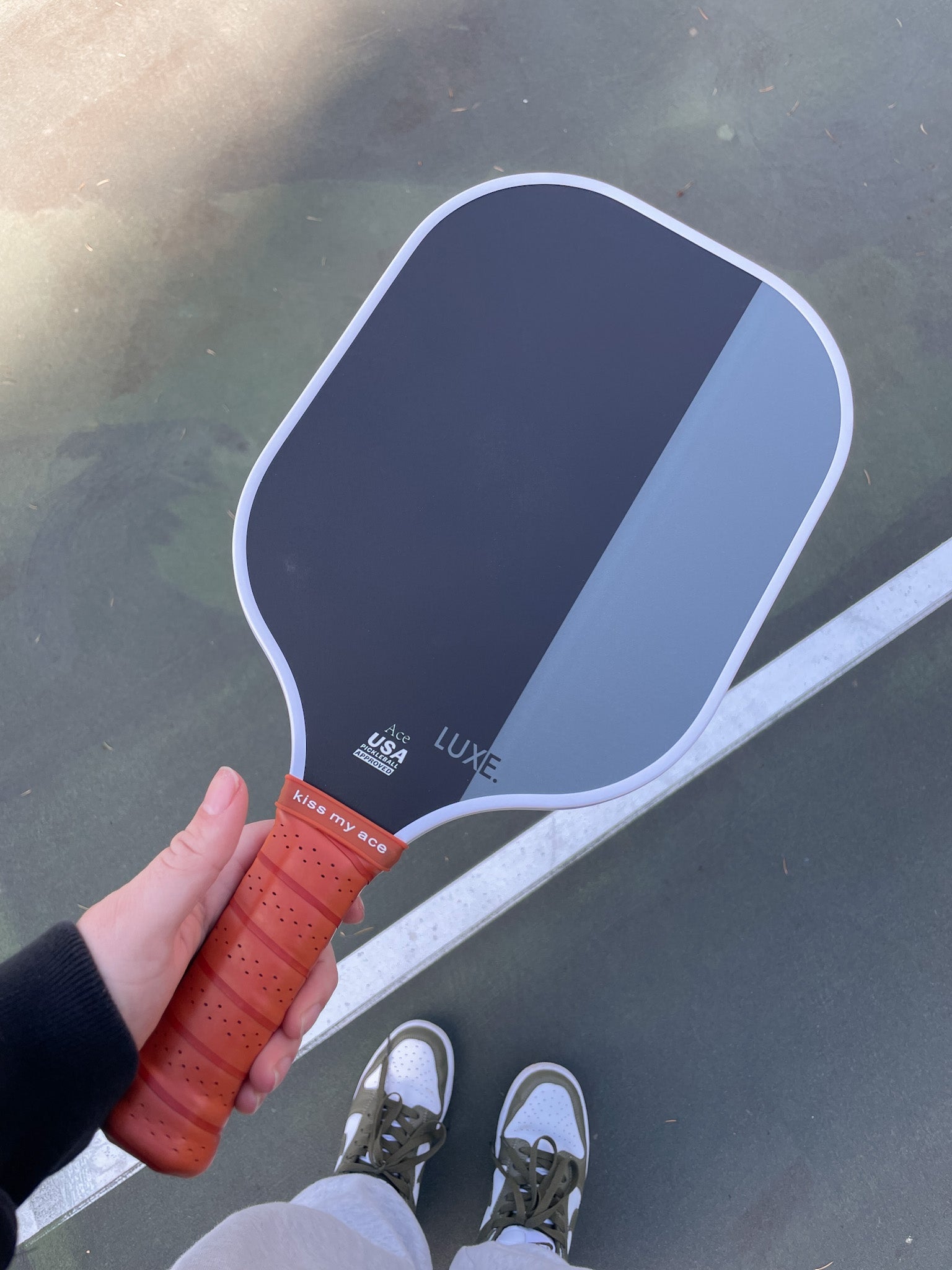 Person holding a Luxe pickleball paddle on a tennis court, wearing white sneakers with green accents.