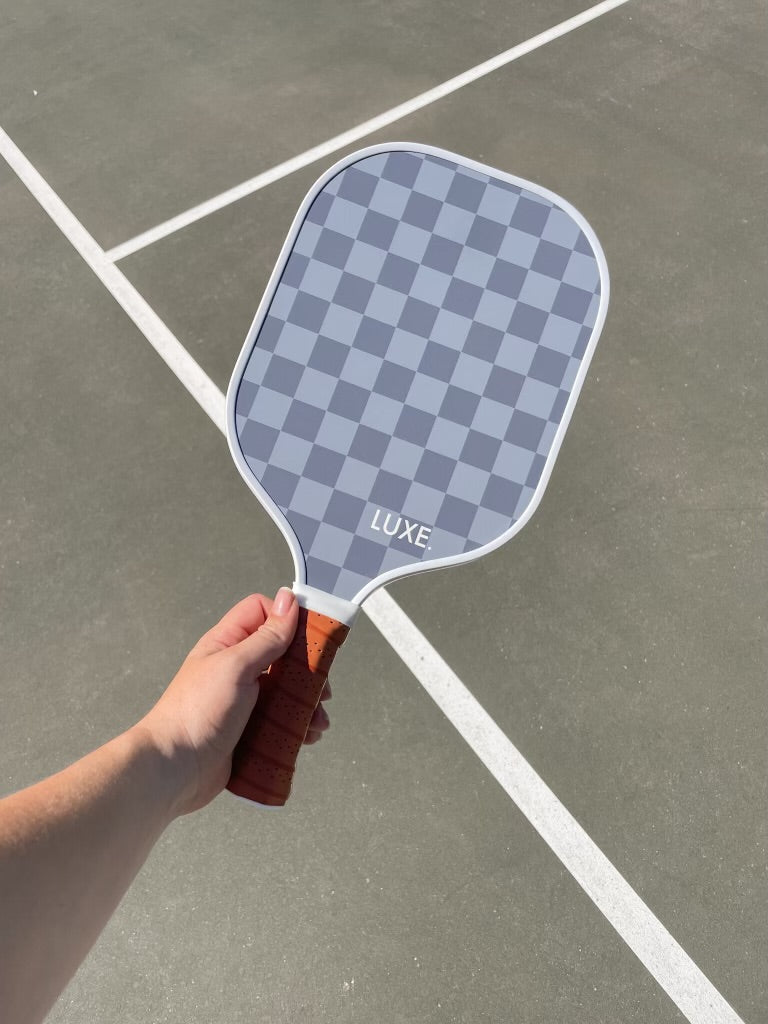 A hand holding a grey and white checkered Luxe pickleball paddle with a brown grip on a grey court with white lines.