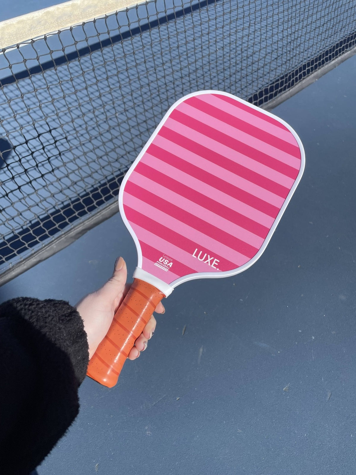 Pink striped pickleball paddle by LUXE Pickleball with an orange grip, held by a hand on a court.