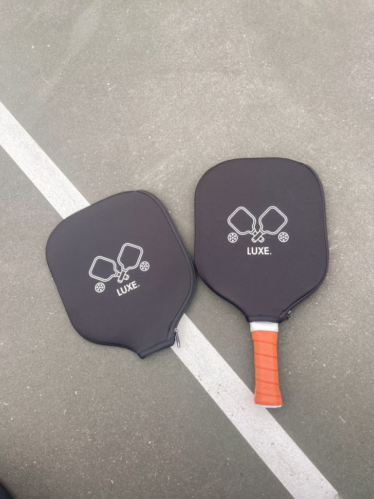 Two black pickleball paddles, one with an orange handle, on a gray court with a white line.