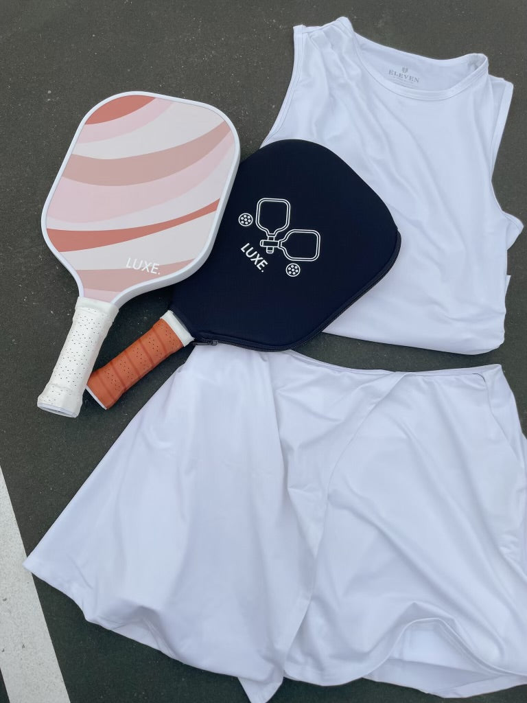 Pickleball paddles, white tank top, and white skirt on a pickleball court.