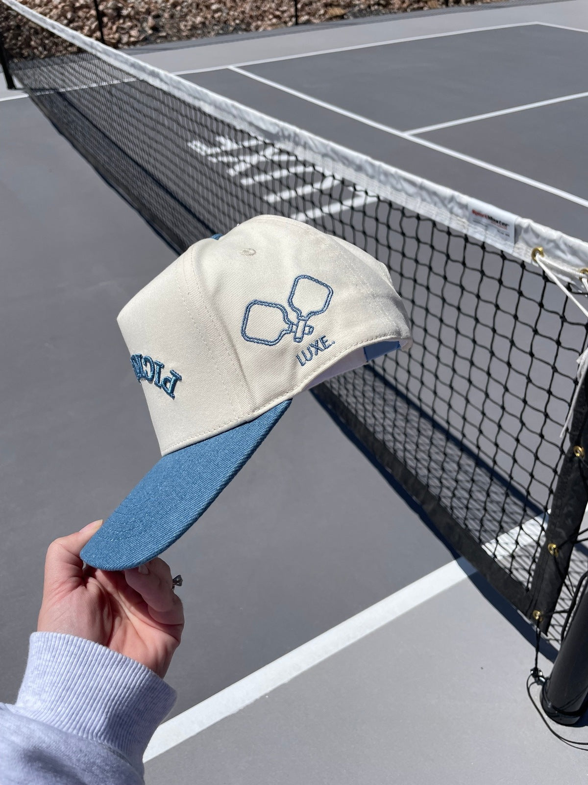 A hand holding a cream and blue hat with pickleball paddles and the word "LUXE" embroidered on it. The background shows a pickleball court.