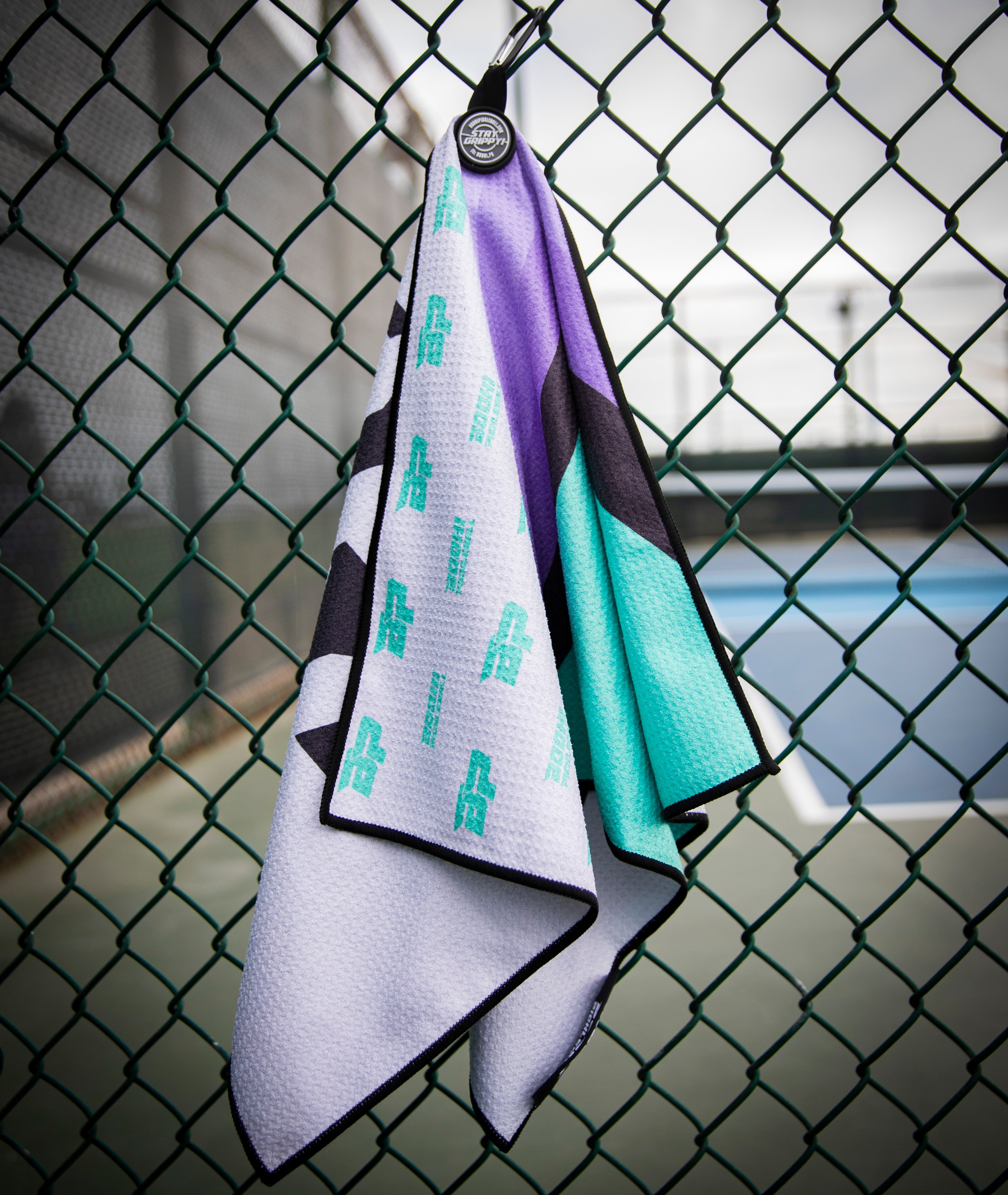 Tennis towel with teal, purple, black, and white colors hanging on a green chainlink fence.