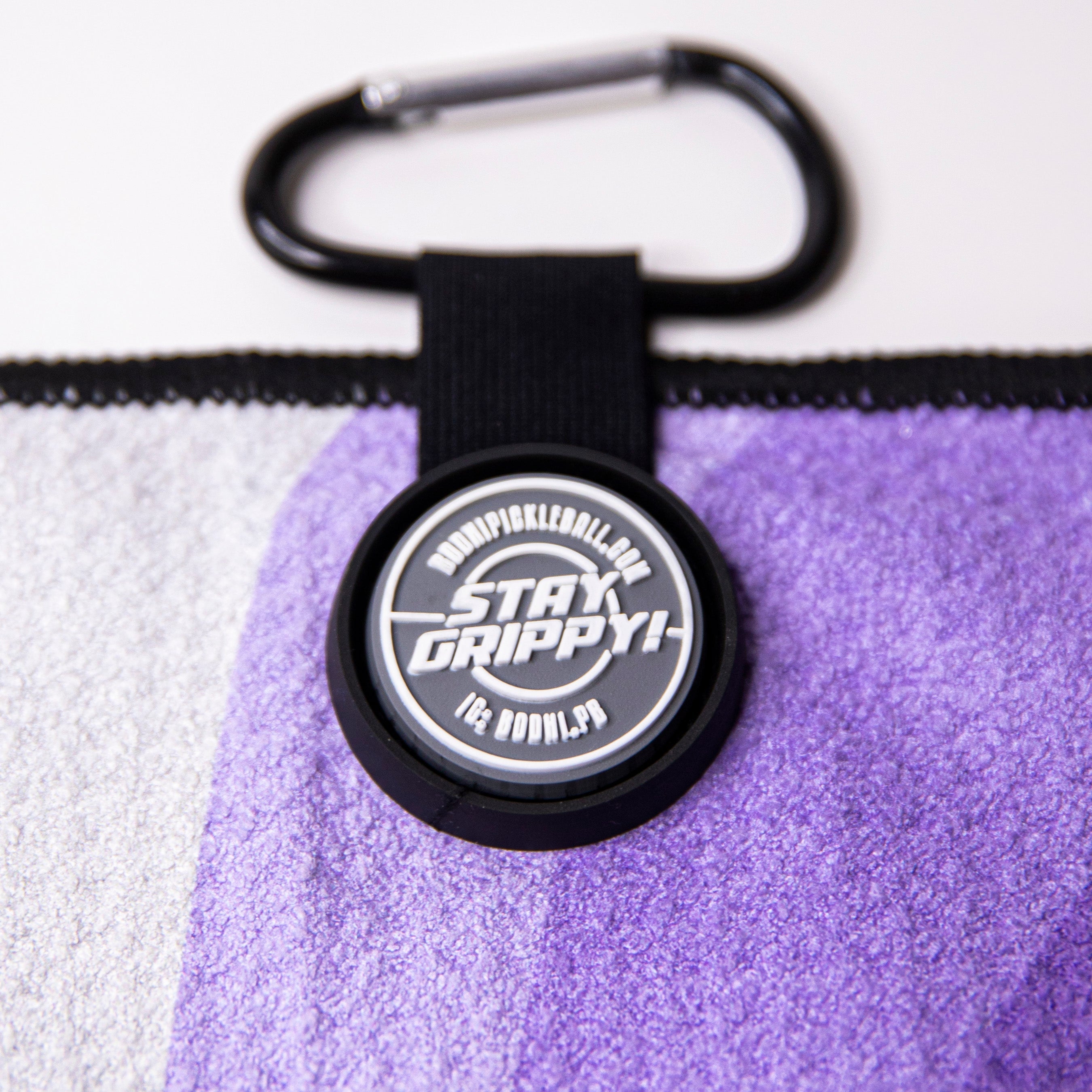 Close-up of a purple pickleball towel with a black carabiner and a round logo that reads "Stay Grippy!".