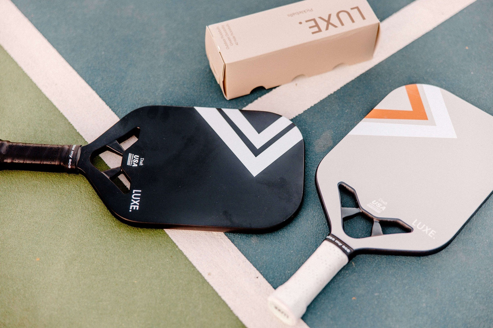 Two pickleball paddles (one black and one gray) and a box of pickleballs on a pickleball court.