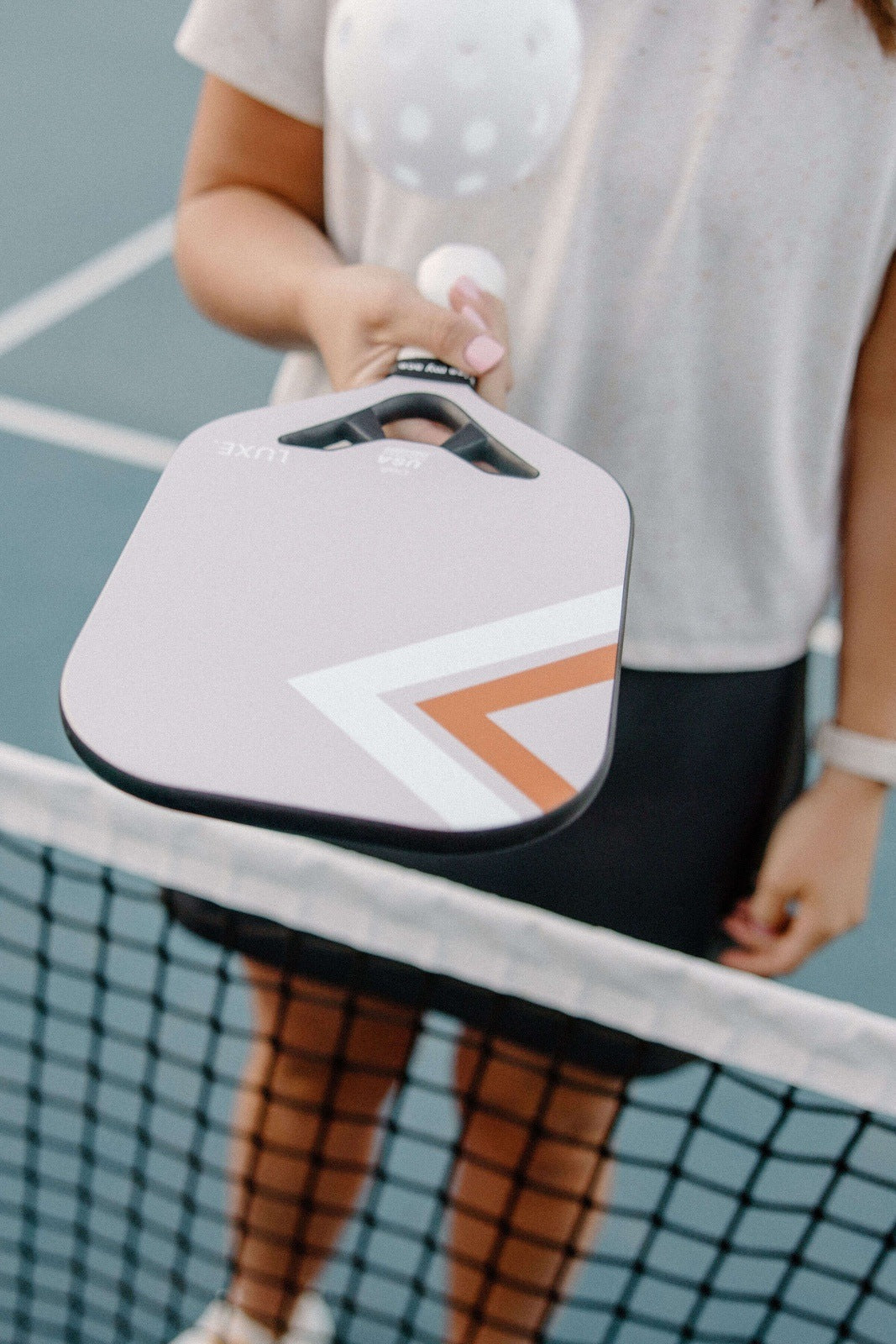 Light gray pickleball paddle with white and orange geometric design by Luxe, held in hand near net on court