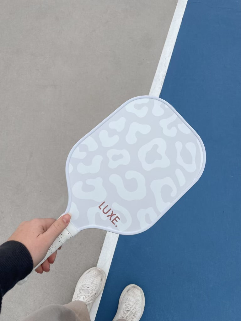Hand holding a LUXE. pickleball paddle with a white and gray cheetah print design, with white sneakers on a blue and gray pickleball court with a white line.