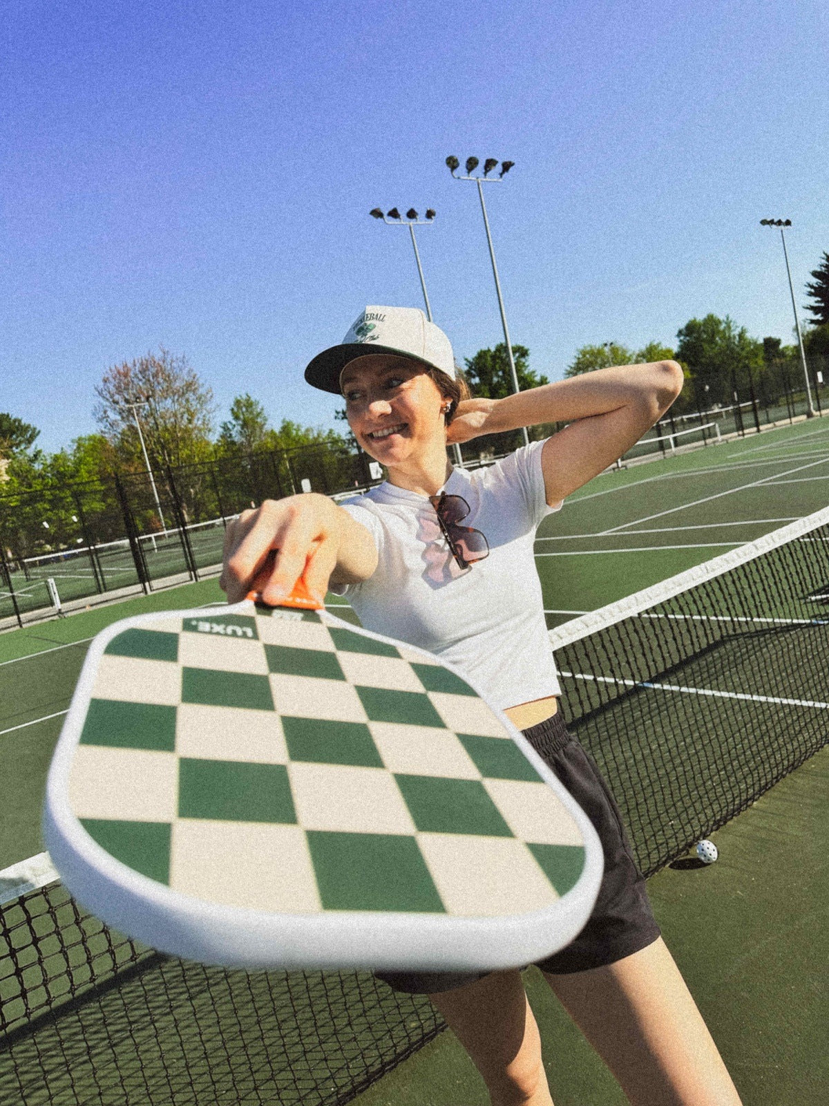 Woman in baseball cap holding checkerboard pickleball paddle on outdoor tennis court.