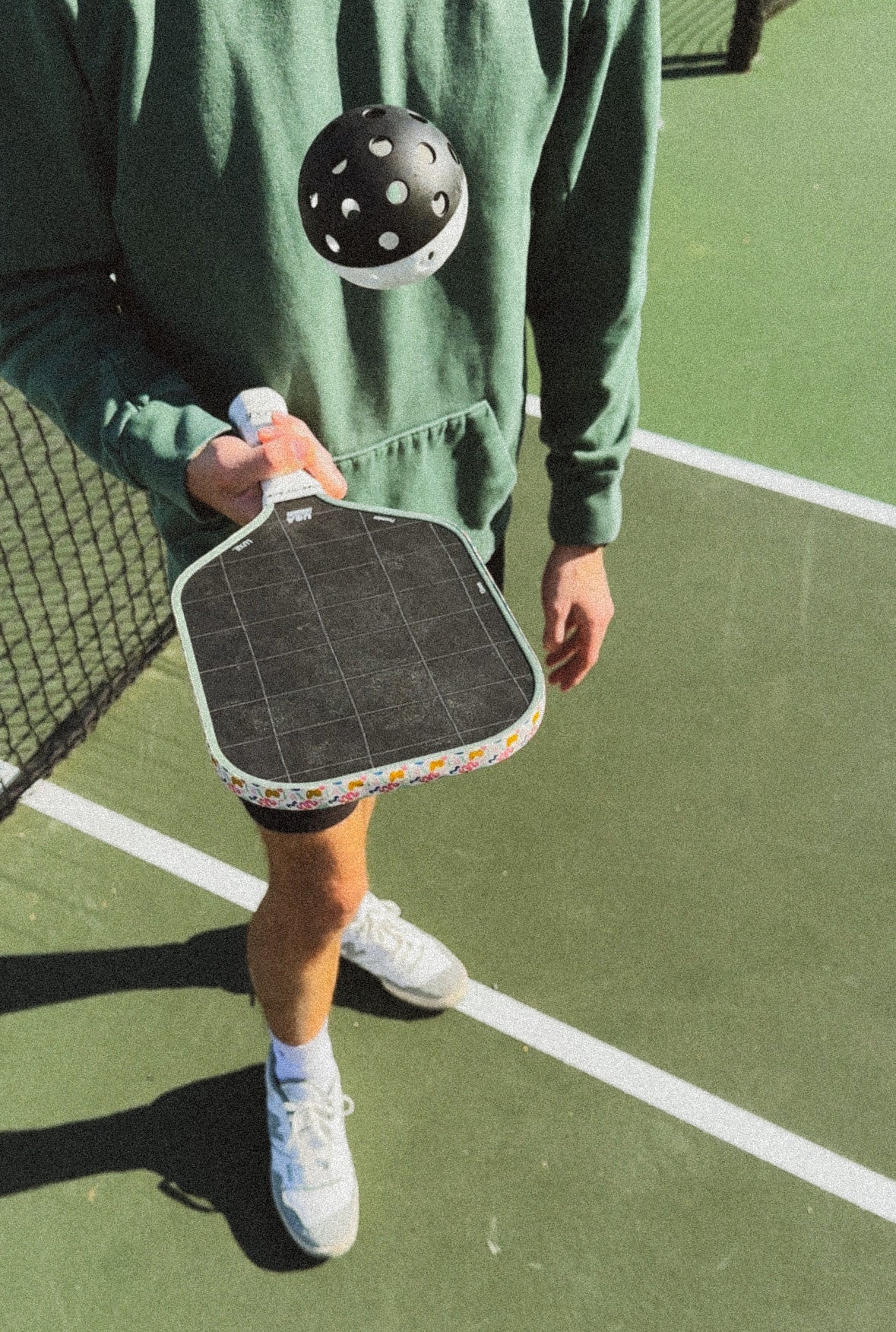 Person in green hoodie holding pickleball paddle and ball on a court.