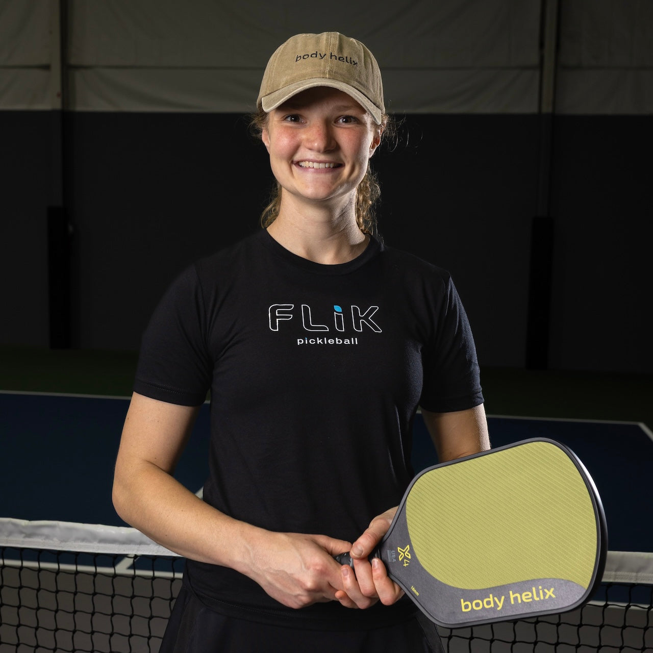 A woman in a black t-shirt and tan hat holds a pickleball paddle.