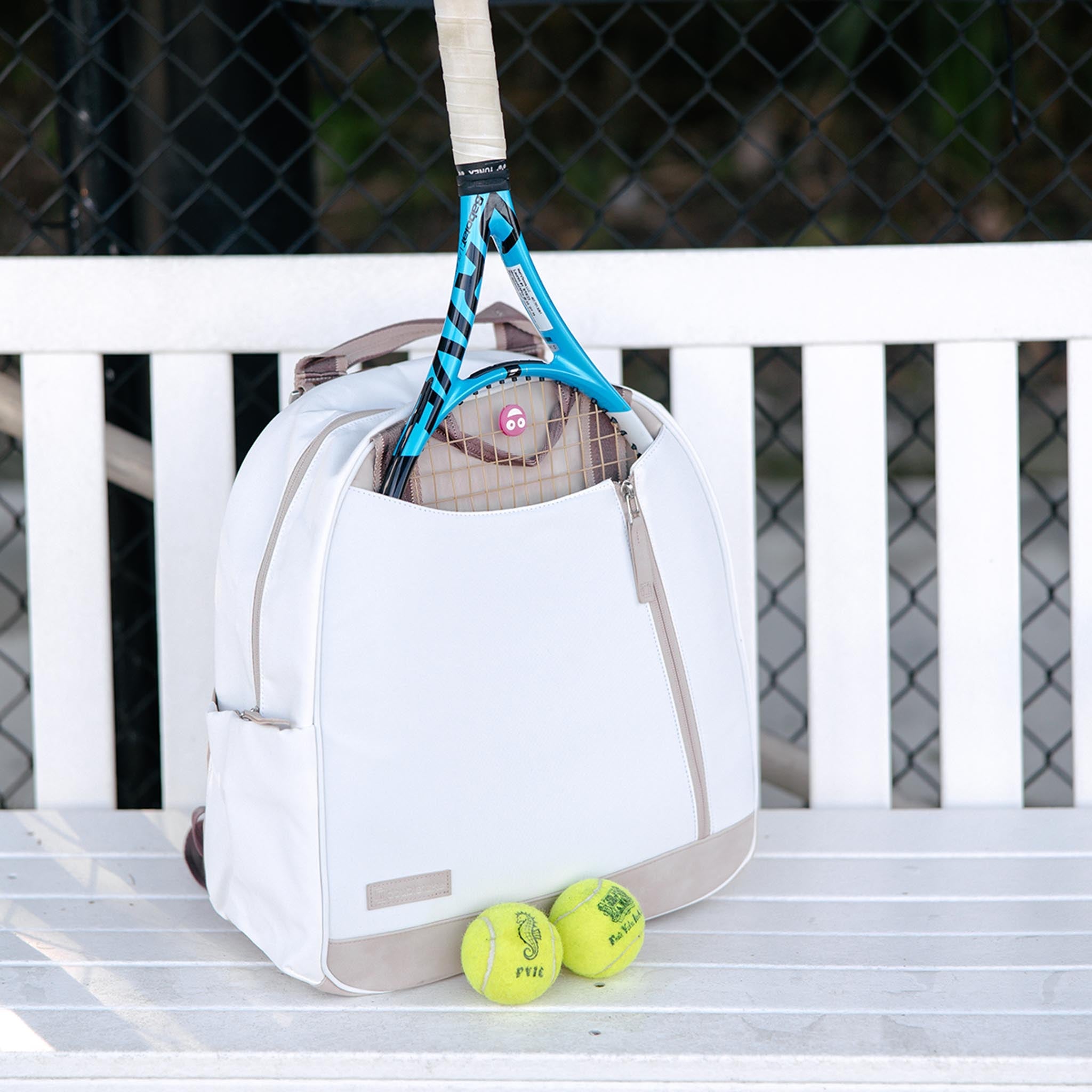 White and beige tennis backpack and tote by Doubletake, holding a racket and two yellow tennis balls on a white slatted bench.