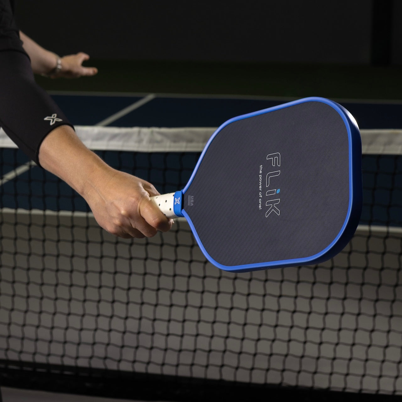Pickleball player holding a "Flik" brand paddle wearing a black arm sleeve, net backdrop.