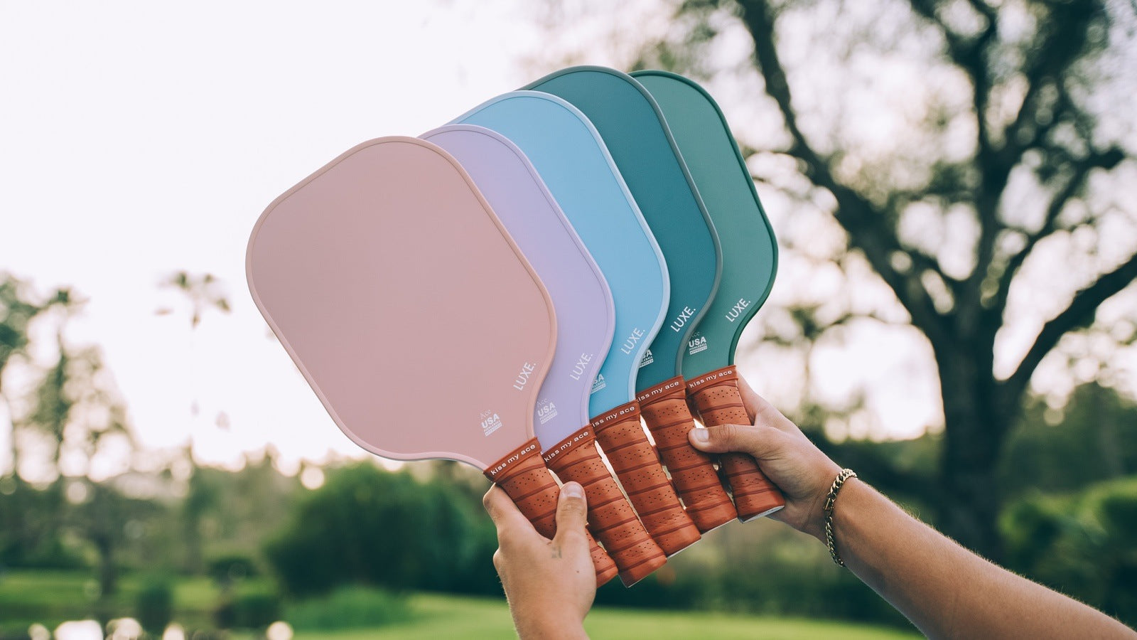A hand holds four colorful LUXE pickleball paddles in front of a blurred outdoor background.
