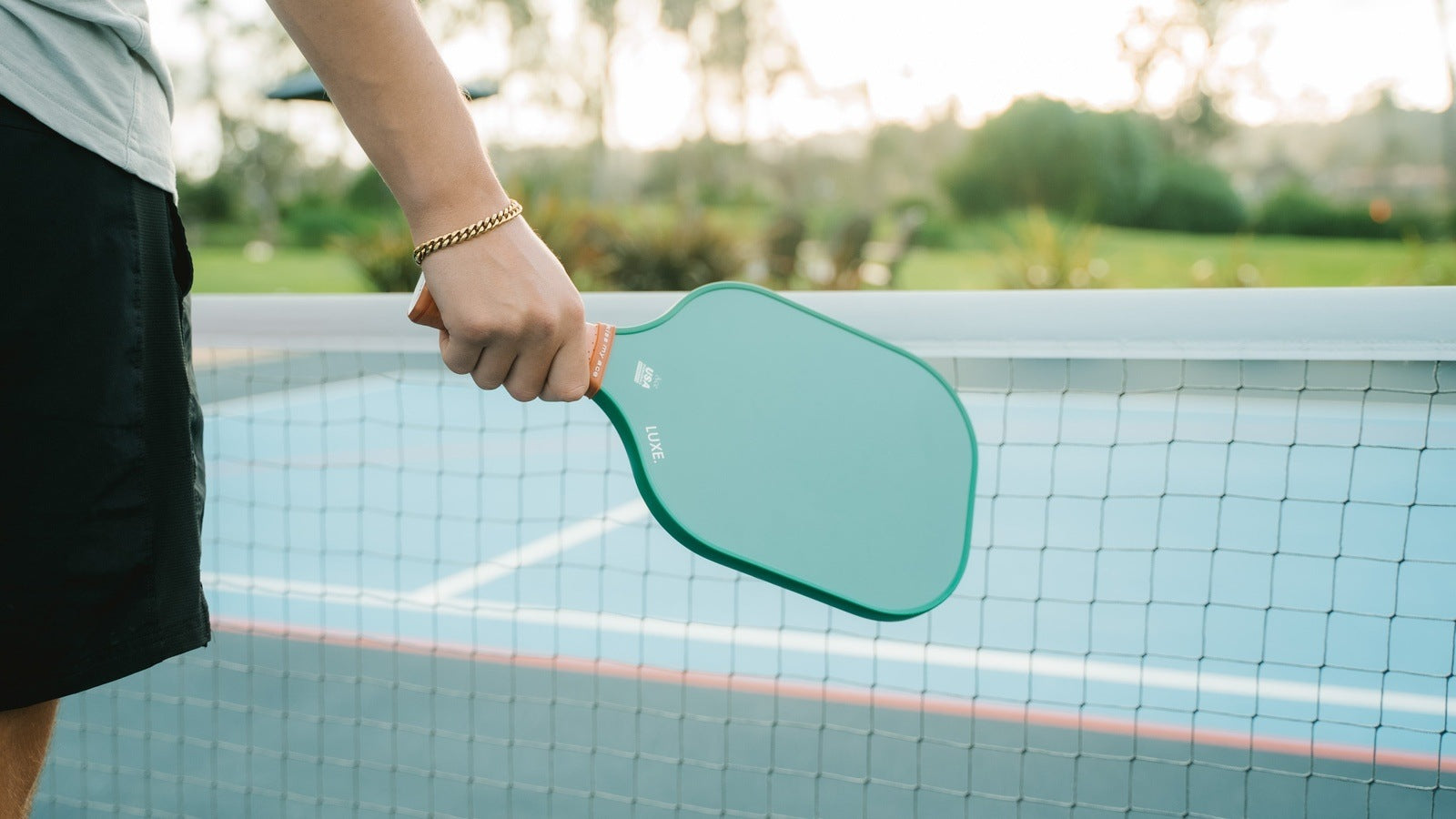 Person holding a turquoise LUXE pickleball paddle on a blue court with net. The person is wearing black shorts and a gold bracelet.