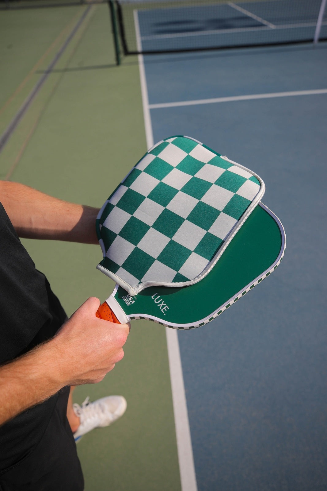 A man holding a green Luxe pickleball paddle with a checkered cover, on a tennis court.
