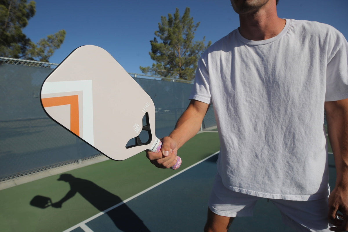 Beige pickleball paddle with orange and white geometric design by Luxe, held in hand on outdoor court, side view, textured grip, shadow visible