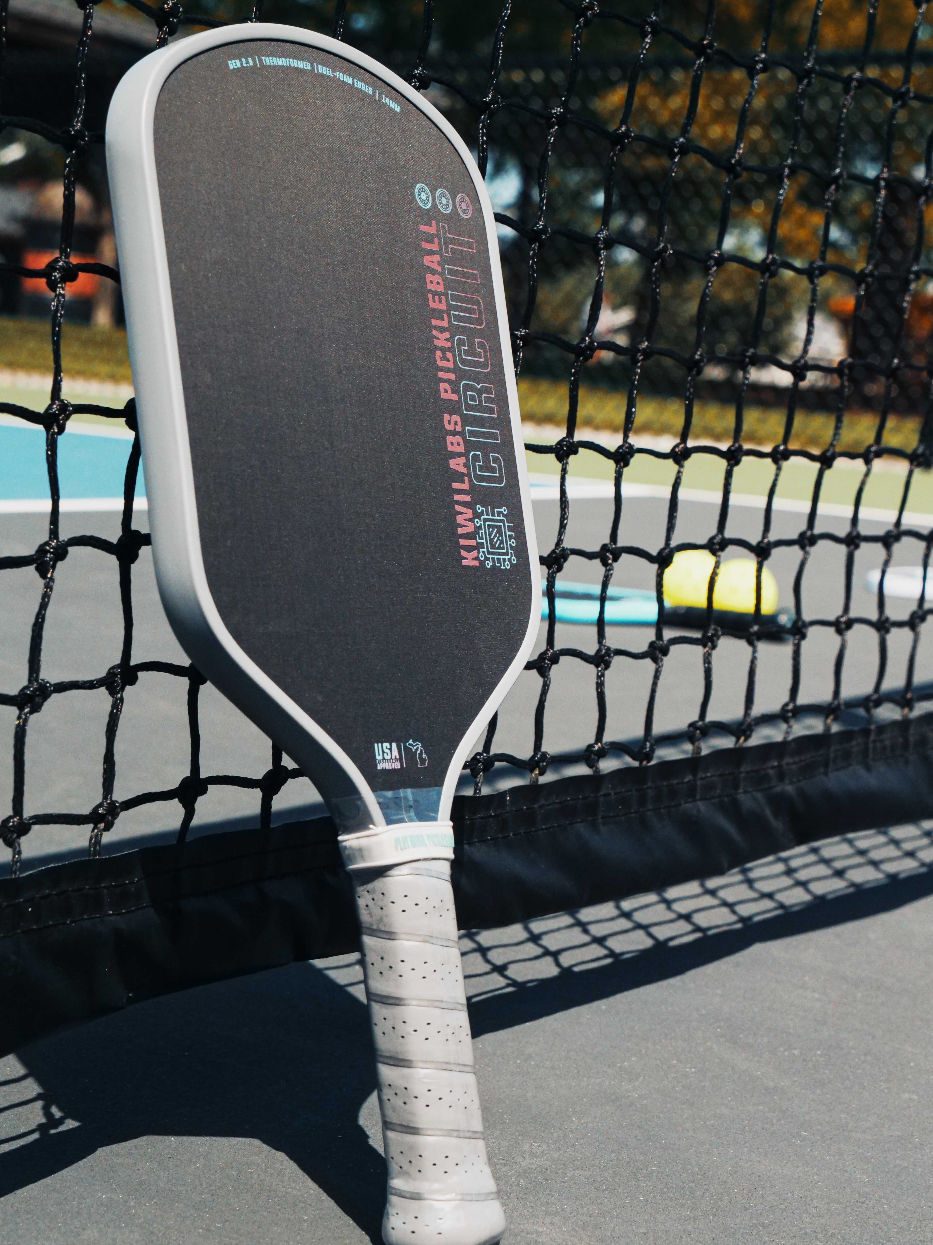 Close-up of a Kiwilabs Circuit pickleball paddle leaning against a net on a court.