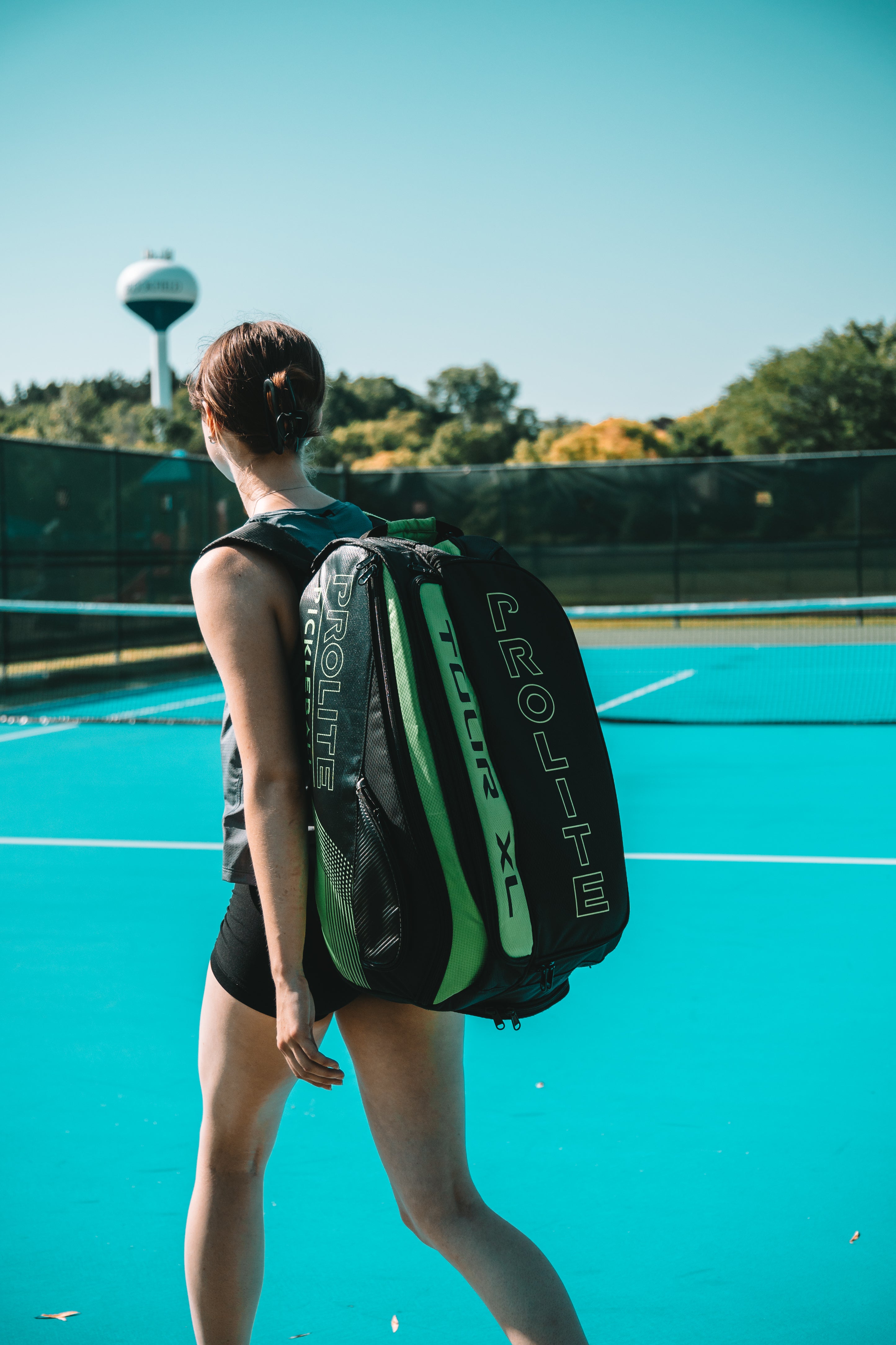 Woman with black and green tennis bag on teal court.