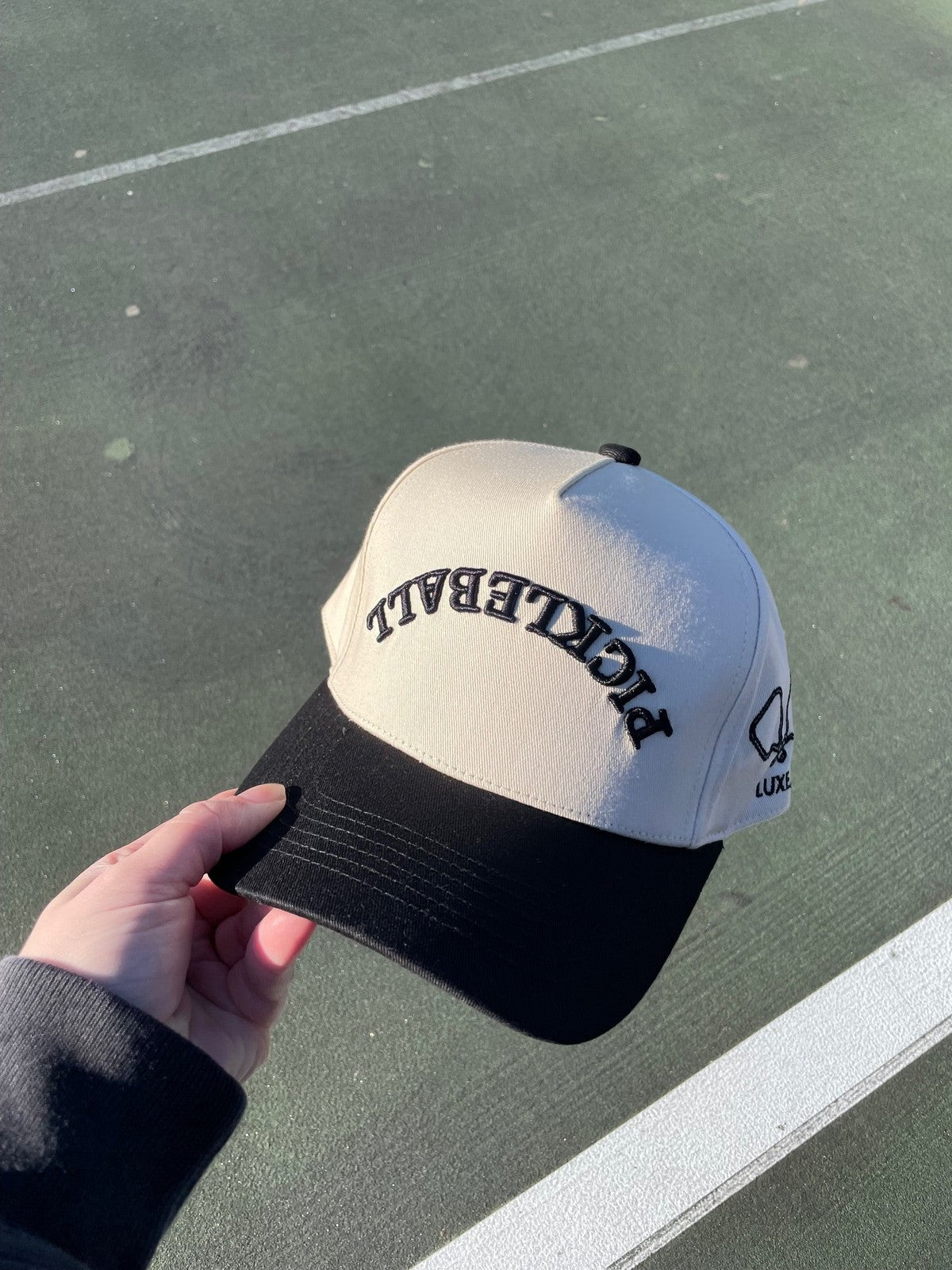 Hand holding a white and black pickleball hat on a green court.