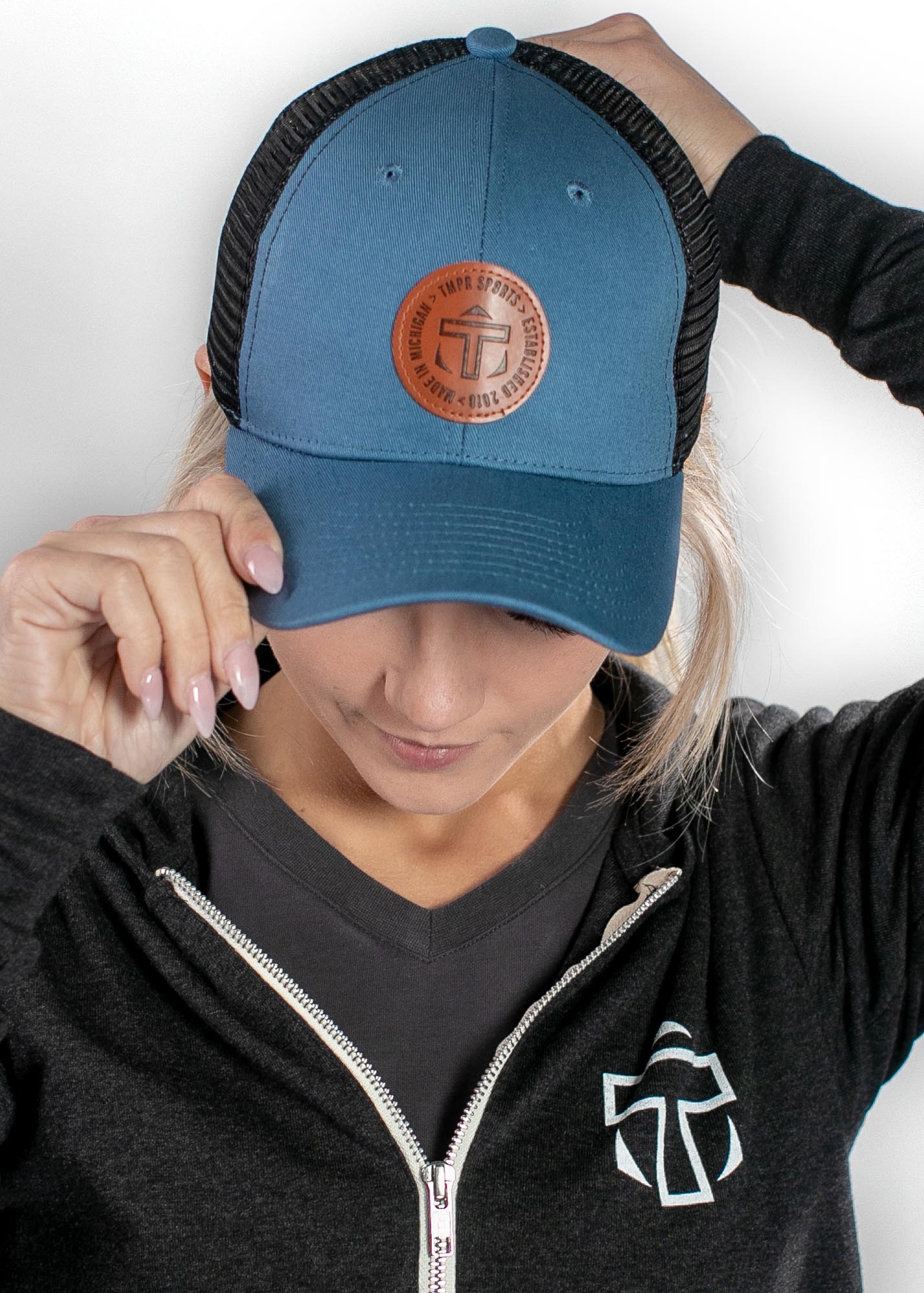 Blue trucker cap by TMPR Sports featuring a brown leather-like logo patch, worn by a woman in a dark grey hoodie.