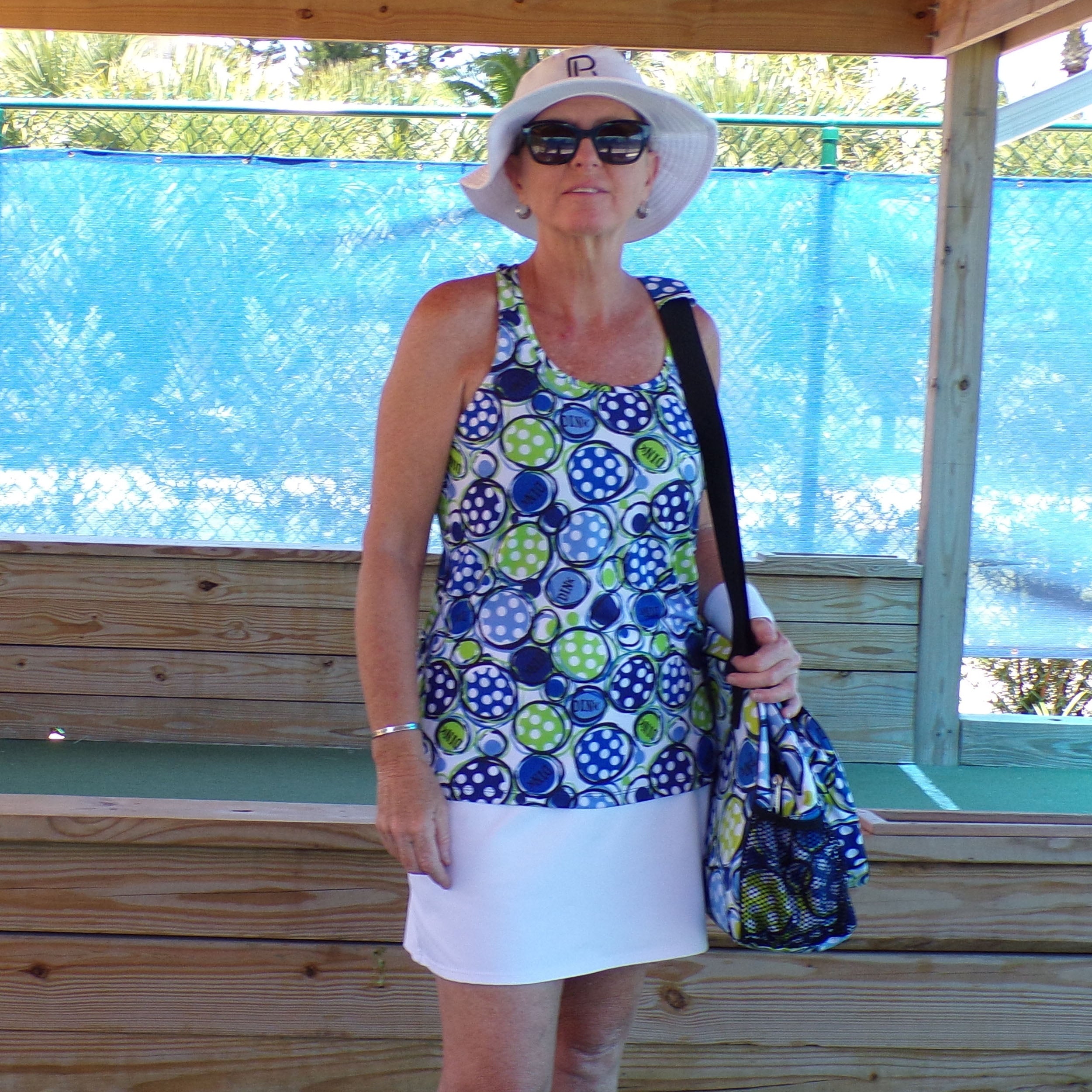 White, blue, and green polka dot racerback tank from Pickleball Bella, featuring 'DINK' text, worn with a white skirt and matching bag.