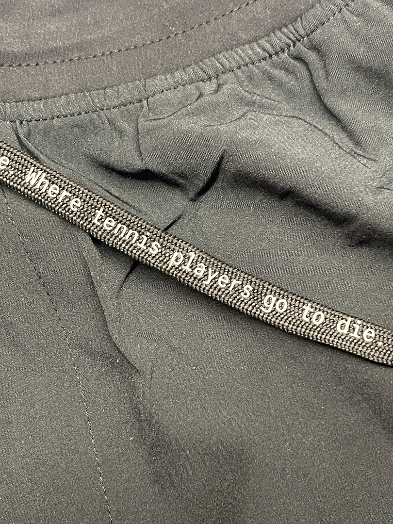 Close-up of black athletic shorts with a drawstring that reads, "Where tennis players go to die".