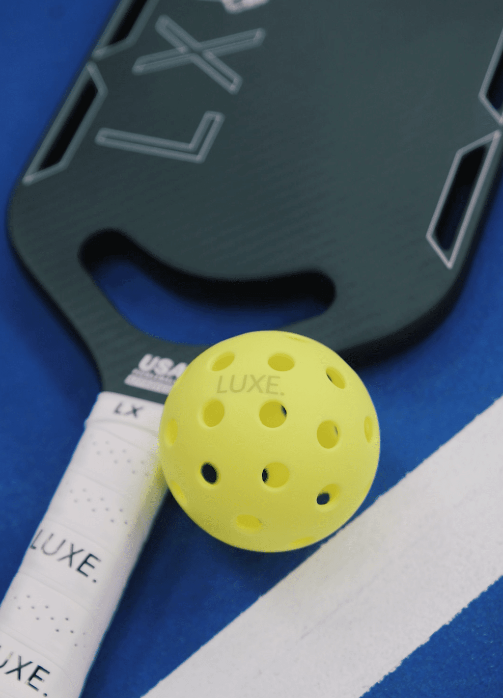 Close-up of a yellow pickleball and green LUXE paddle on a blue surface with a white line.