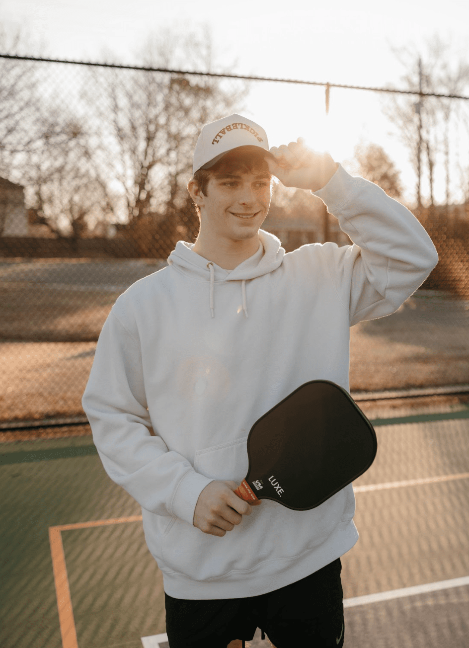 Young man in a white hoodie and "Pickleball" hat, holding a black pickleball paddle, on a pickleball court at sunset.