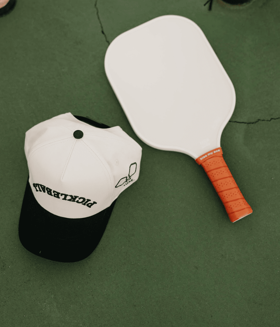 A white and black pickleball cap and white paddle with orange grip on a green surface.