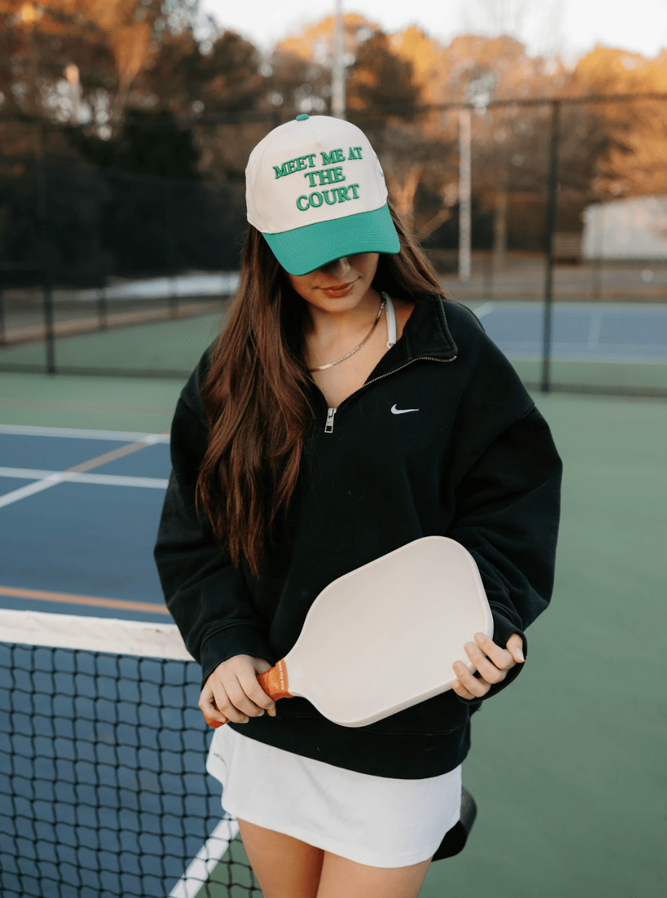 A woman holding a pickleball paddle wears a hat that says "Meet Me At The Court" and a black Nike sweatshirt.