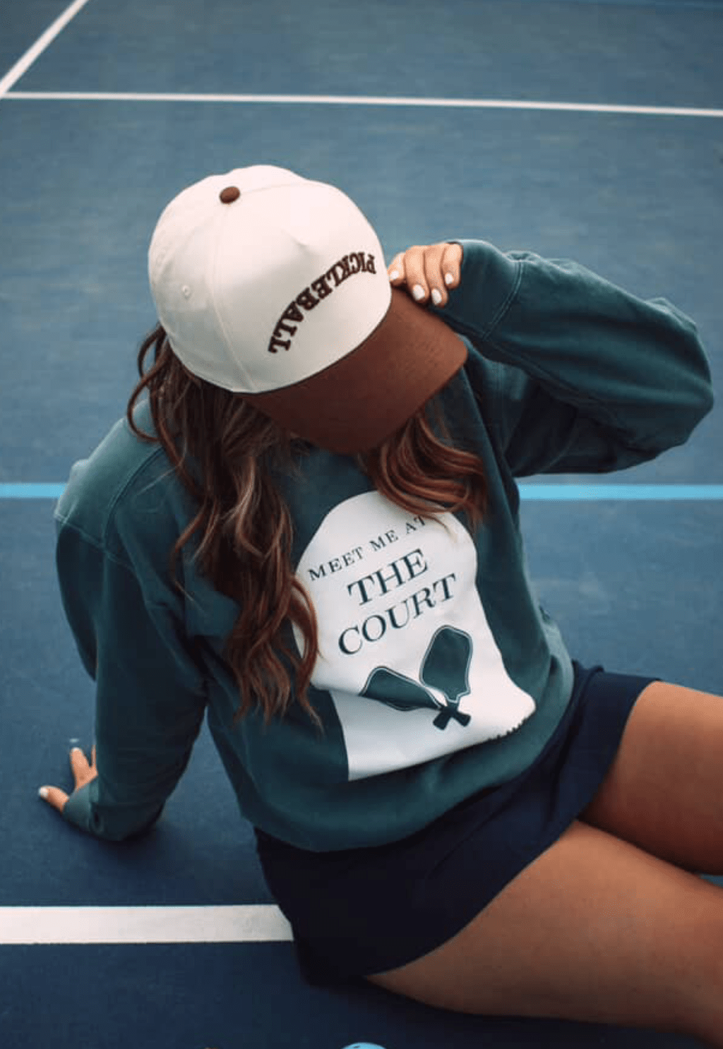 Woman in a white and brown pickleball hat, teal sweatshirt with "Meet me at the court" printed on it, and blue skirt sitting on a blue court.