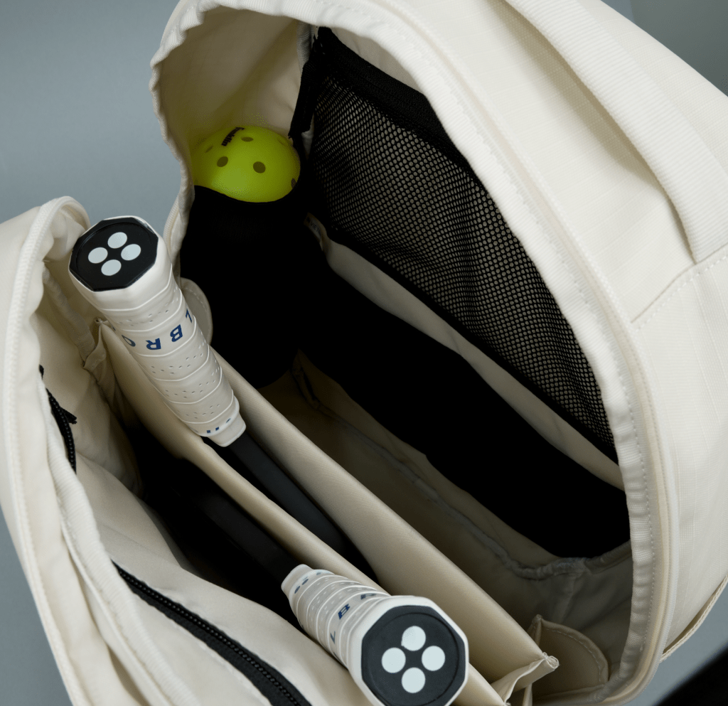 Cream pickleball backpack by FORWRD, open from overhead, revealing organized compartments, two white-gripped paddles, and a yellow pickleball.