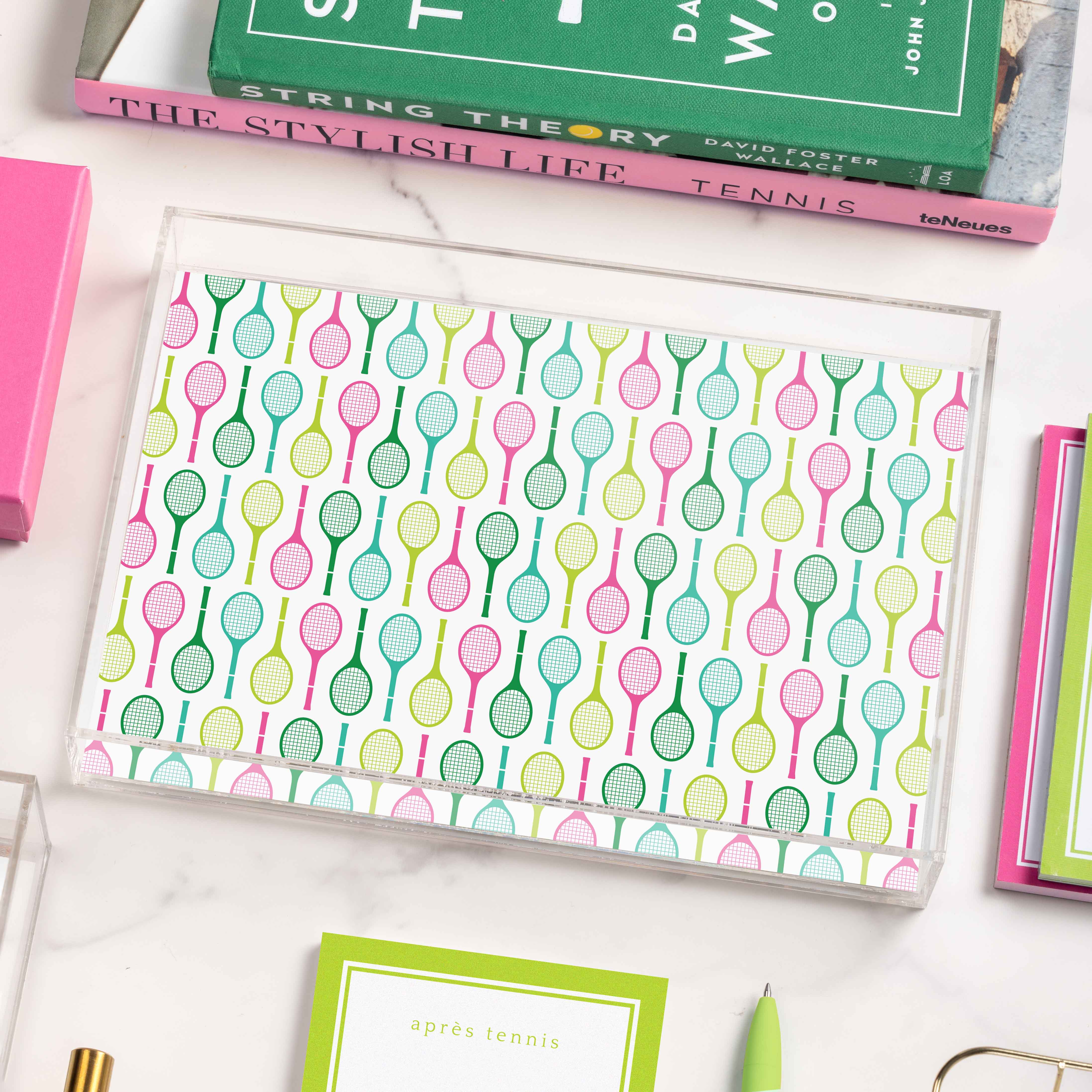An acrylic tray with a tennis racquet pattern rests on a white marble surface, surrounded by books and stationery in bright pink, green, and yellow.