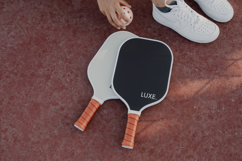 Close-up of pickleball paddles with orange grip, a pickleball in a hand, and white sneakers on a reddish textured surface.