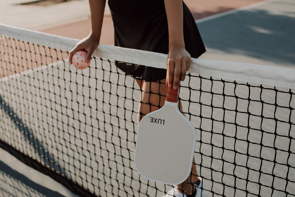 Woman in black skirt standing at a pickleball net holding a paddle and ball.