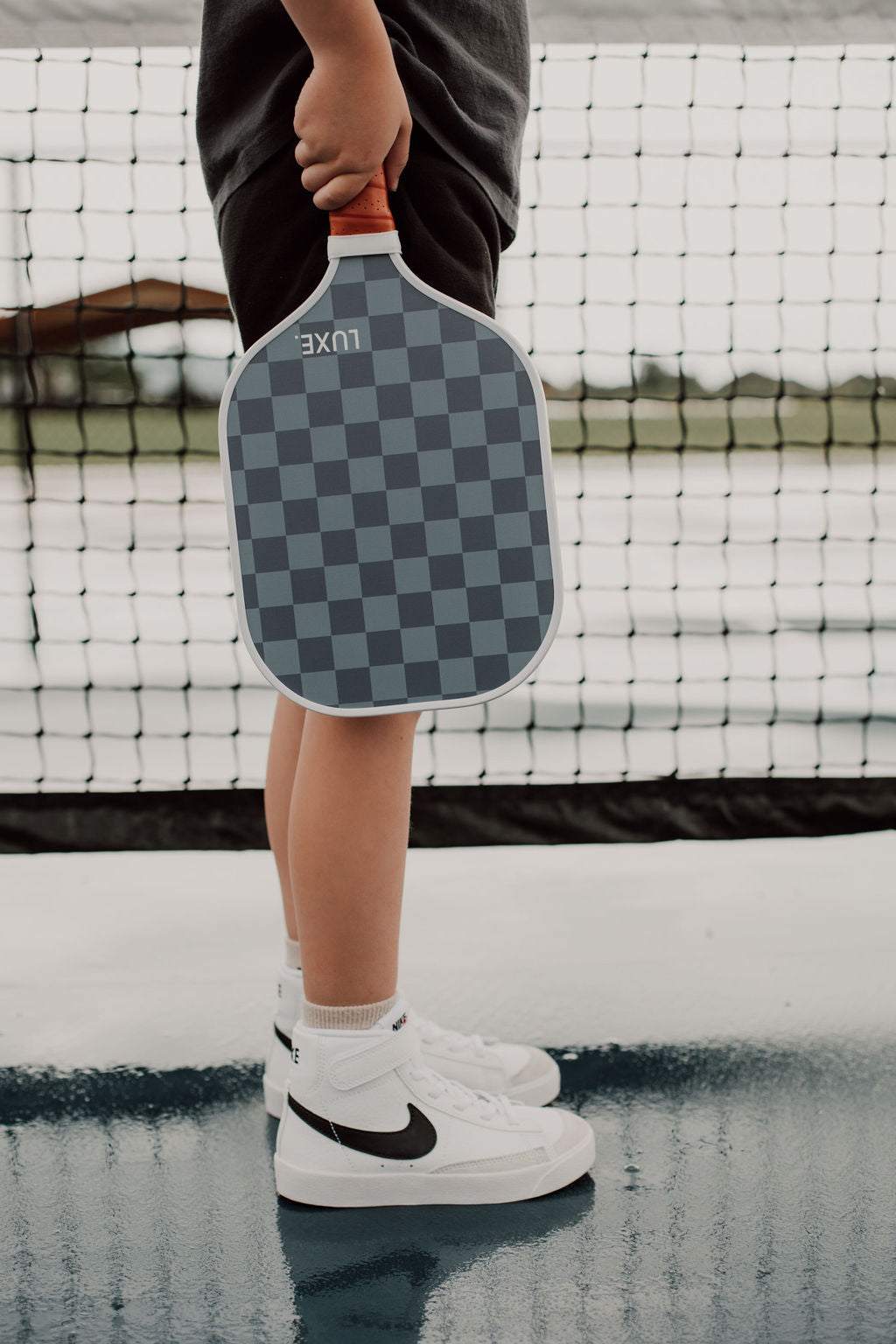 A person holding a checkerboard pickleball paddle in front of a net, wearing white Nike high-top sneakers.