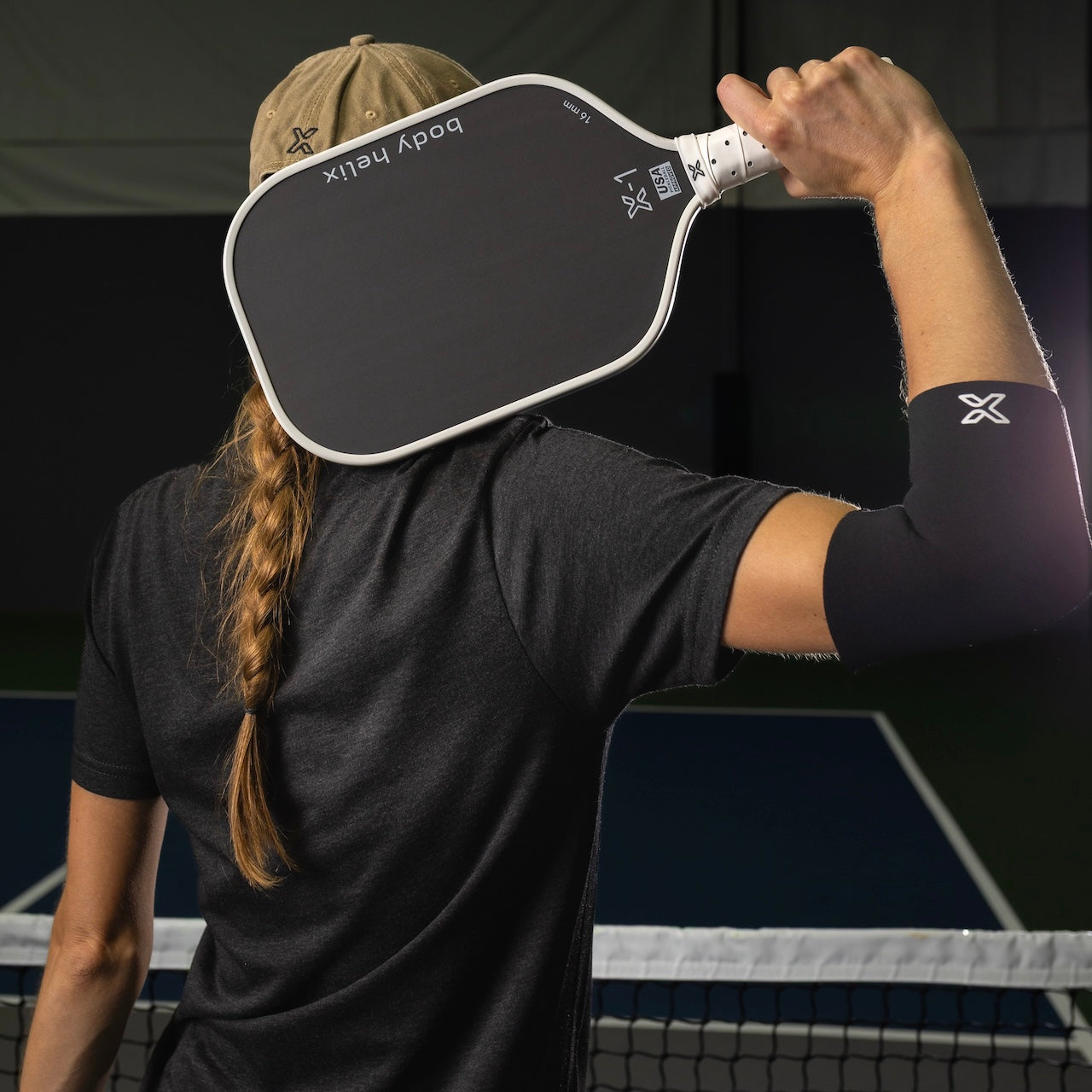 A woman in a gray t-shirt and tan hat holding a black and white pickleball paddle.