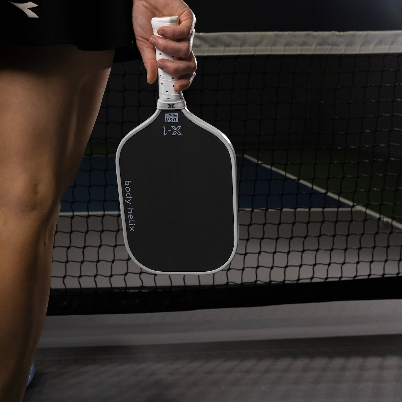 Woman holding a black Body Helix X-1 pickleball paddle with a white handle in front of a pickleball net.