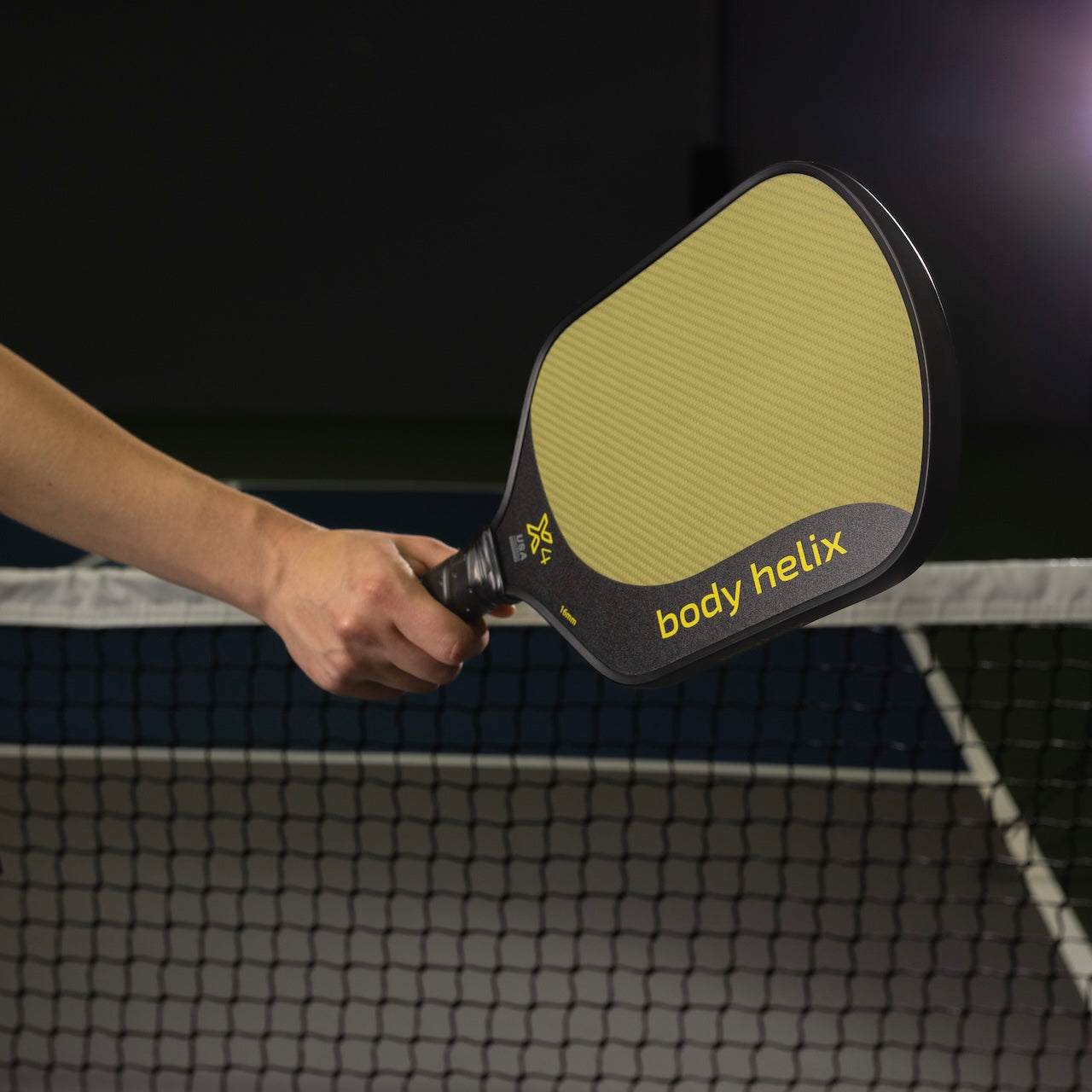 A hand holding a yellow Body Helix pickleball paddle in front of a pickleball net.